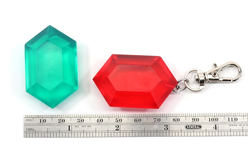 May include: Two plastic Triforce keychains, one green and one red. The red keychain has a silver metal clasp. A ruler is in the bottom of the image to show the size of the keychains.
