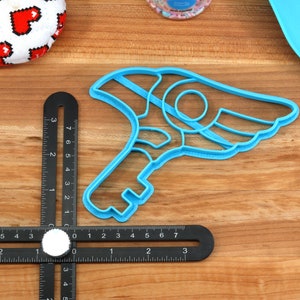 May include: A blue plastic cookie cutter shaped like a key with a wing on the side. The cutter is on a wooden surface with a ruler.