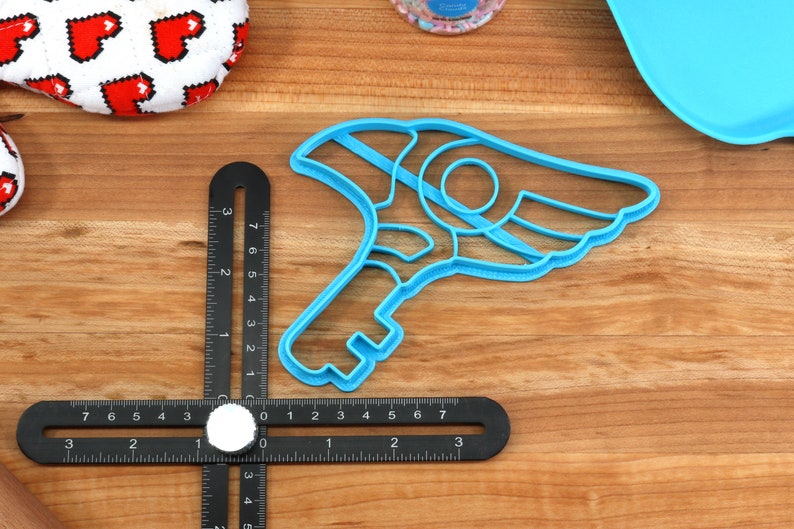 May include: A blue plastic cookie cutter shaped like a key with a wing on the side. The cutter is on a wooden surface with a ruler.