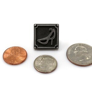 May include: A silver pin with a black and white image of a high heel shoe. The pin is surrounded by three US coins: a quarter, a dime, and a penny.