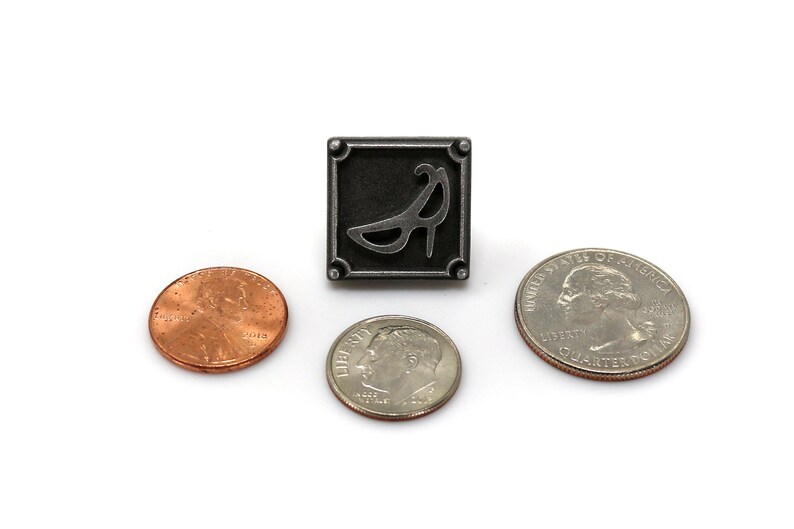 May include: A silver pin with a black and white image of a high heel shoe. The pin is surrounded by three US coins: a quarter, a dime, and a penny.