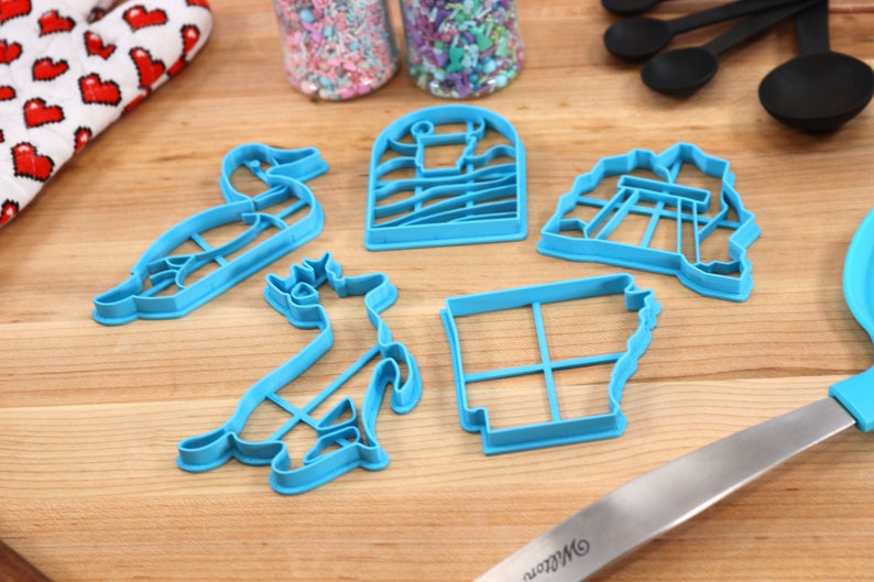 Op de afbeelding: Een set van vijf blauwe plastic koekjesvormen in de vorm van een eend, een hert, een vuurtoren, een brug en de staat Arkansas.
