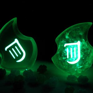 Op de afbeelding: Twee groene, gloeiende harsversieringen met een wit muzieknotensymbool. De versieringen zijn vlamvormig.