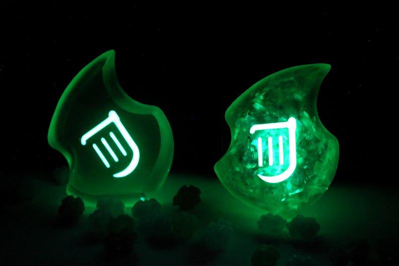Op de afbeelding: Twee groene, gloeiende harsversieringen met een wit muzieknotensymbool. De versieringen zijn vlamvormig.