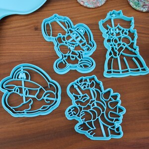 May include: Four blue plastic cookie cutters shaped like characters from the Super Mario Bros. video game franchise. The cutters are on a wooden surface.