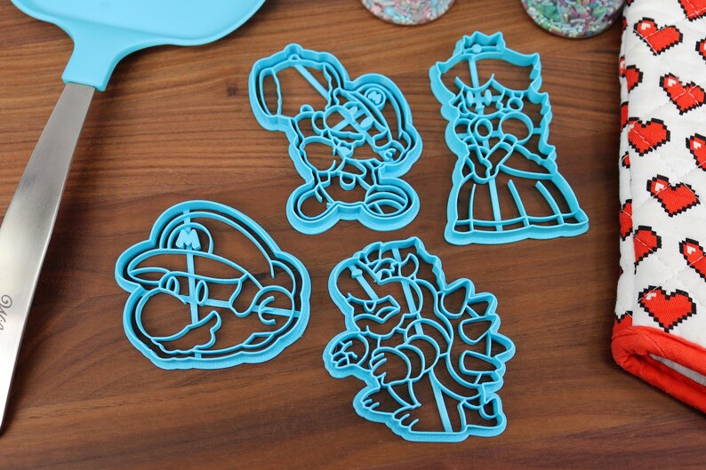 May include: Four blue plastic cookie cutters shaped like characters from the Super Mario Bros. video game franchise. The cutters are on a wooden surface.