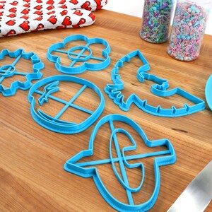 May include: A collection of blue plastic cookie cutters in various shapes, including a gear, flower, and a horse. A blue spatula and jars of sprinkles are also present on a wooden cutting board. A red and white heart-patterned oven mitt is in the background.