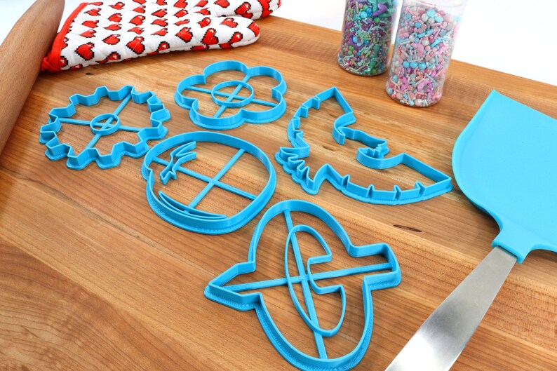 May include: A collection of blue plastic cookie cutters in various shapes, including a gear, flower, and a horse. A blue spatula and jars of sprinkles are also present on a wooden cutting board. A red and white heart-patterned oven mitt is in the background.