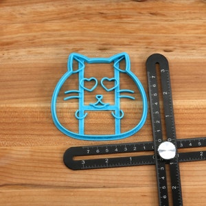 May include: Blue plastic cookie cutter shaped like a cat with heart-shaped eyes. The cutter is on a wooden surface with a black ruler.
