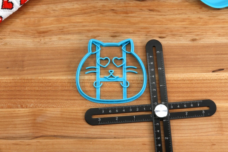 May include: Blue plastic cookie cutter shaped like a cat with heart-shaped eyes. The cutter is on a wooden surface with a black ruler.