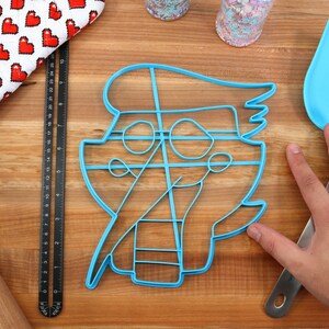 May include: A blue plastic cookie cutter shaped like a cartoon pirate with a hat, eye patch, and a sword. The cutter is on a wooden surface.