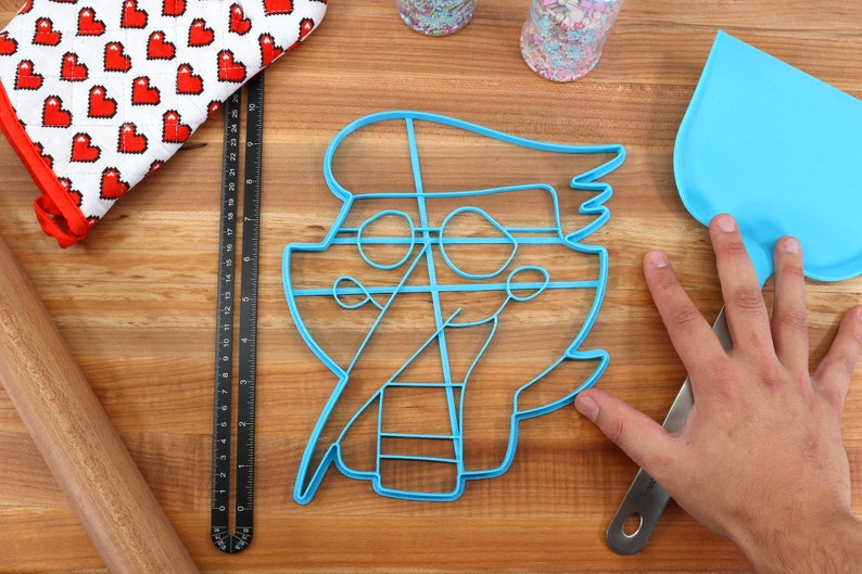 May include: A blue plastic cookie cutter shaped like a cartoon pirate with a hat, eye patch, and a sword. The cutter is on a wooden surface.