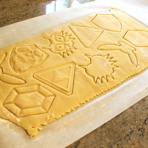 Puede incluir: Una hoja de masa para galletas con varias formas cortadas, incluyendo una espada, un escudo, un coraz&oacute;n, una estrella y un diamante. Las formas est&aacute;n todas cortadas de la masa y listas para ser horneadas.