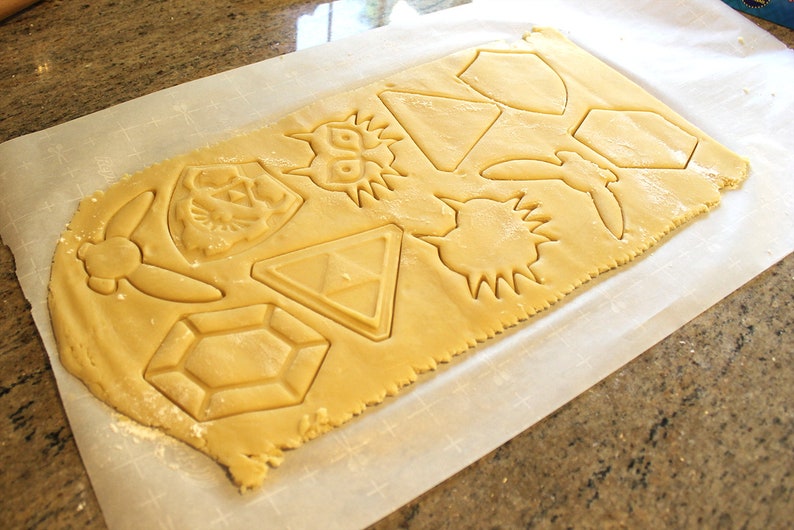 Puede incluir: Una hoja de masa para galletas con varias formas cortadas, incluyendo una espada, un escudo, un coraz&oacute;n, una estrella y un diamante. Las formas est&aacute;n todas cortadas de la masa y listas para ser horneadas.