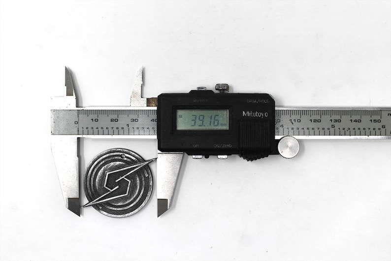 May include: A silver metal pin with a stylized lightning bolt design is measured with a digital caliper. The caliper reads 39.16 mm.