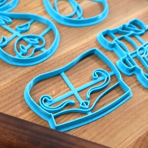 May include: Several light blue plastic cookie cutters in various shapes, including a bow, a piano, and other decorative designs. The cutters are arranged on a wooden surface.