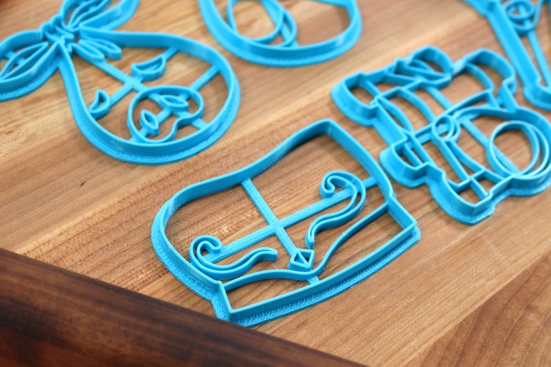 May include: Several light blue plastic cookie cutters in various shapes, including a bow, a piano, and other decorative designs. The cutters are arranged on a wooden surface.