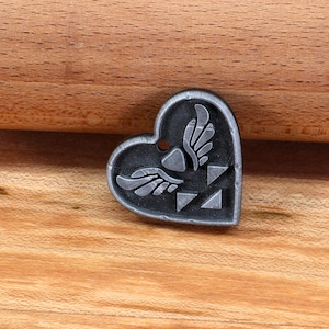 Puede incluir: Un amuleto de plata en forma de coraz&oacute;n con un dise&ntilde;o alado estilizado y un patr&oacute;n triangular. El amuleto est&aacute; sobre una superficie de madera.