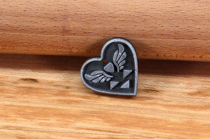 Puede incluir: Un amuleto de plata en forma de coraz&oacute;n con un dise&ntilde;o alado estilizado y un patr&oacute;n triangular. El amuleto est&aacute; sobre una superficie de madera.