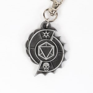 May include: A silver metal keychain featuring a stylized dice with a skull and a sun symbol. The keychain is attached to a silver metal ring.