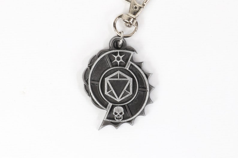 May include: A silver metal keychain featuring a stylized dice with a skull and a sun symbol. The keychain is attached to a silver metal ring.