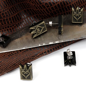 May include: Black and gold-colored earrings with a crown and heart design. The earrings are displayed on a metal surface and a brown textured background. The earrings have a post-back closure.