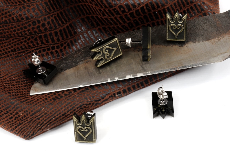May include: Black and gold-colored earrings with a crown and heart design. The earrings are displayed on a metal surface and a brown textured background. The earrings have a post-back closure.