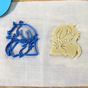 May include: Two cookie cutters, one blue and one beige, shaped like a cartoon character with short hair and a mask over their mouth and nose.