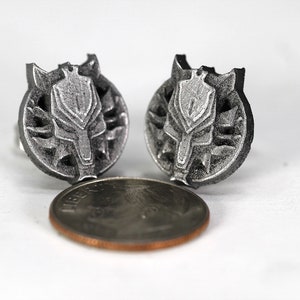 Op de afbeelding: Een paar zilverkleurige metalen oorbellen met een wolfskopontwerp. De oorbellen zijn ongeveer zo groot als een Nederlandse cent.