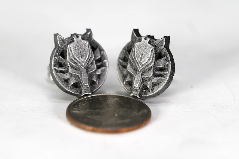 Op de afbeelding: Een paar zilverkleurige metalen oorbellen met een wolfskopontwerp. De oorbellen zijn ongeveer zo groot als een Nederlandse cent.