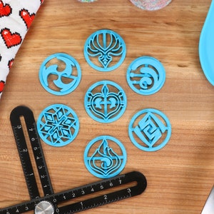 May include: Eight blue plastic cookie cutters with different designs, including a snowflake, a heart, a flame, and a swirl. The cutters are on a wooden surface with a black ruler and a blue spatula.