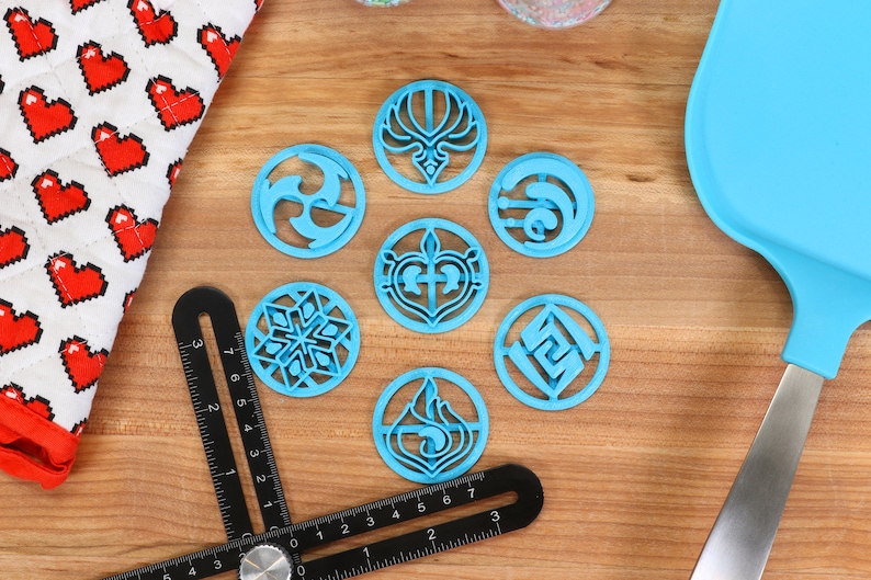 May include: Eight blue plastic cookie cutters with different designs, including a snowflake, a heart, a flame, and a swirl. The cutters are on a wooden surface with a black ruler and a blue spatula.