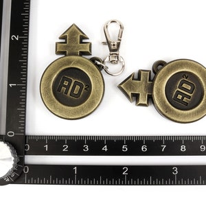 May include: Two gold-colored metal keychains with a circular design featuring the text "AD&sup2;" and a stylized arrow symbol. The keychain on the left has a silver metal clasp.