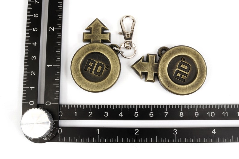 May include: Two gold-colored metal keychains with a circular design featuring the text "AD&sup2;" and a stylized arrow symbol. The keychain on the left has a silver metal clasp.