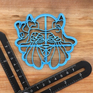 May include: A blue plastic cookie cutter shaped like a cartoon character with large eyes, a wide mouth, and horns. The character is wearing a hat with a cross on top.