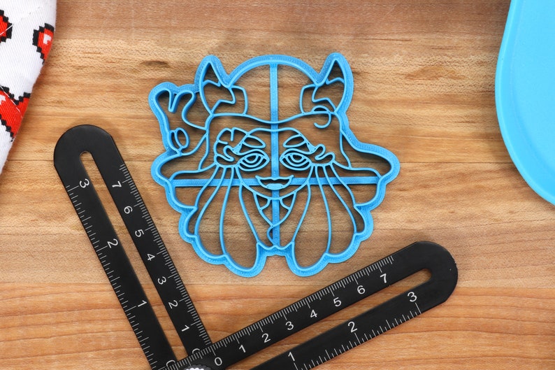 May include: A blue plastic cookie cutter shaped like a cartoon character with large eyes, a wide mouth, and horns. The character is wearing a hat with a cross on top.
