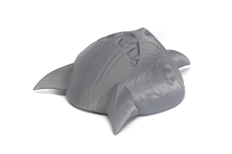 May include: A gray 3D printed shark head with a toothy grin and detailed fins. The shark head is a decorative object.