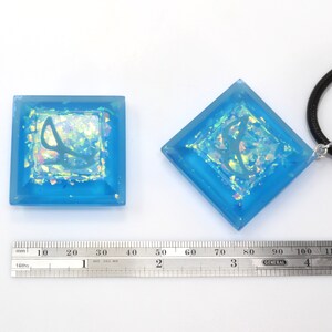 May include: Two blue square resin pendants with iridescent glitter and a silver design. One pendant is on a black cord necklace. A ruler is in the image for size reference.