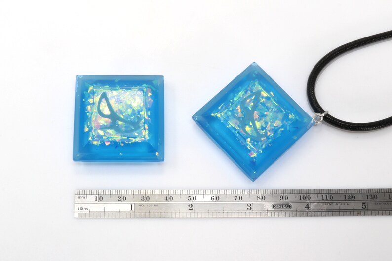 May include: Two blue square resin pendants with iridescent glitter and a silver design. One pendant is on a black cord necklace. A ruler is in the image for size reference.