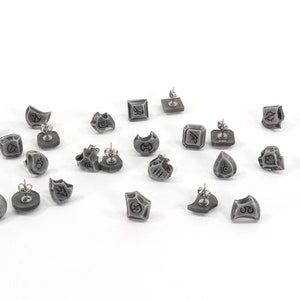 May include: A collection of gray stud earrings with various geometric and symbolic designs. Each earring has a silver post. The earrings are scattered on a white surface, showcasing their unique shapes and details.
