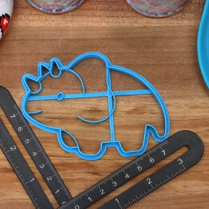 May include: A blue plastic cookie cutter shaped like a triceratops dinosaur. The cutter is on a wooden surface with a ruler and a blue silicone mat.