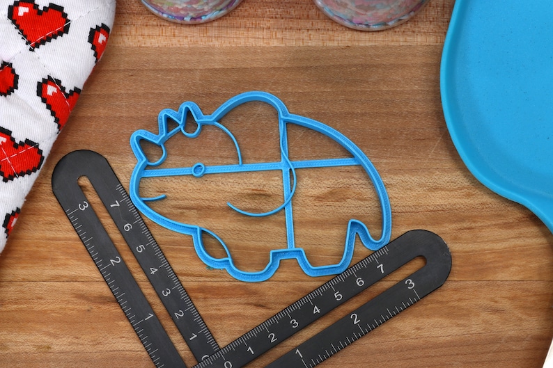 May include: A blue plastic cookie cutter shaped like a triceratops dinosaur. The cutter is on a wooden surface with a ruler and a blue silicone mat.