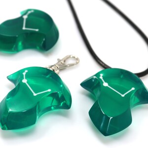 May include: A collection of green resin pendants and charms. The pendants are shaped like stylized figures with a white constellation design. One pendant is on a black cord, another has a silver clasp, and a third is a standalone charm.