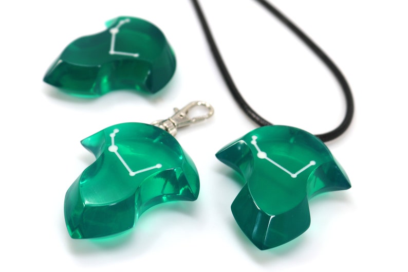 May include: A collection of green resin pendants and charms. The pendants are shaped like stylized figures with a white constellation design. One pendant is on a black cord, another has a silver clasp, and a third is a standalone charm.