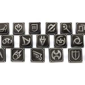 May include: A set of 18 silver-toned metal pins featuring various symbols, including a heart, a musical note, a sword, and a shield. The pins are arranged in rows on a white background.