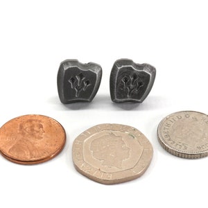 以下が含まれることがあります： スタイリッシュな花柄のデザインが施された盾型のシルバーメタル製のイヤリングのペア。イヤリングは、スケールのために米国の1セント硬貨と英国の1ポンド硬貨の隣に置かれています。