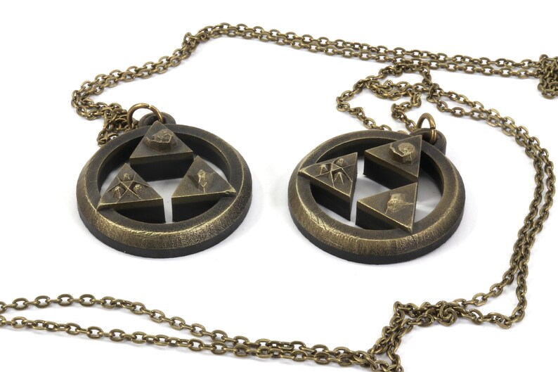 Puede incluir: Dos collares de color dorado con un colgante circular que presenta un dise&ntilde;o de estrella de tres puntas. Cada colgante tiene un dise&ntilde;o diferente en cada punta de la estrella.