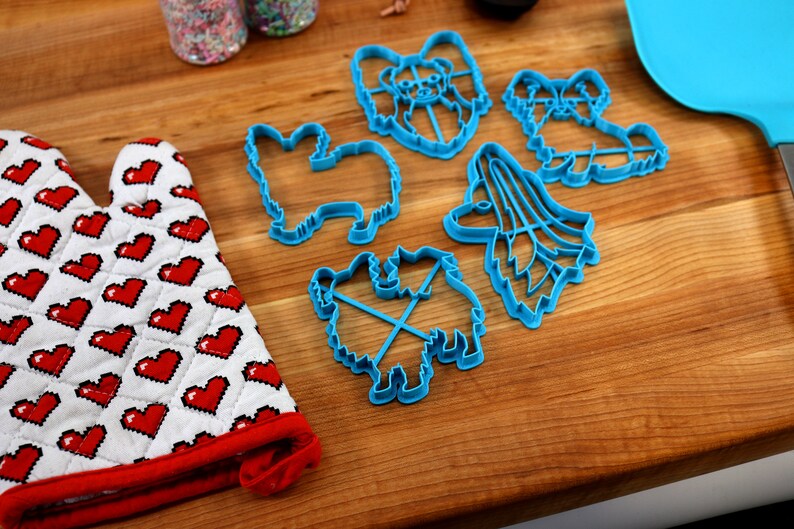 May include: A set of five blue plastic cookie cutters shaped like different breeds of dogs. The cutters are on a wooden surface.