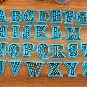 May include: A set of 26 blue plastic cookie cutters shaped like the letters of the alphabet. Each letter has a decorative design around the edge.
