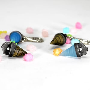 May include: Three different drill bit keychains, one blue, one brown, and one gray, with silver metal accents. The keychains are scattered on a white surface with small colorful candies.